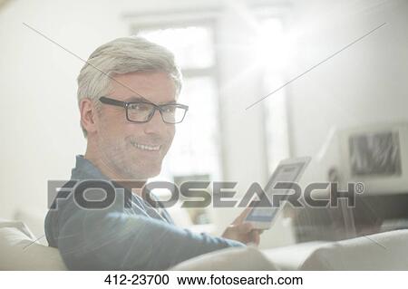 Stock Image - Older man using digital tablet on living room sofa. Fotosearch