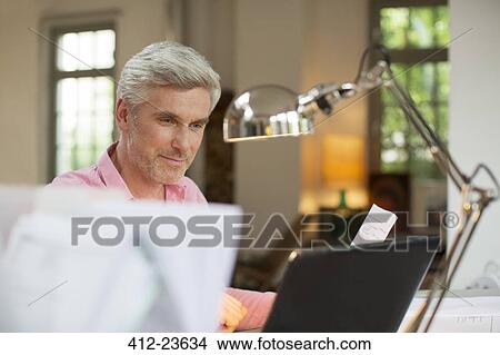 Picture - Older man using laptop in home office. Fotosearch