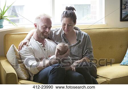 Stock Image - Parents smiling and holding little baby, sitting on yellow sofa. Fotosearch
