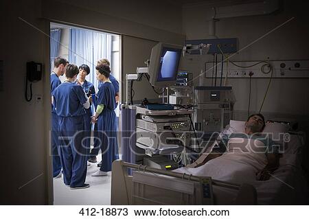 Stock Image - Patient lying in bed in intensive care unit, team of doctors discussing in background. Fotosearch