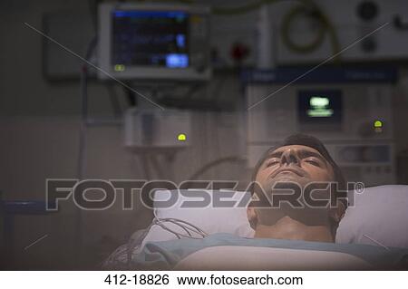 Stock Photograph - Patient lying in bed, surrounded by monitoring medical equipment in intensive care unit. Fotosearch