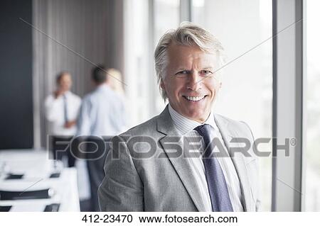 Stock Image - Portrait of businessman standing in conference room with colleagues in background. Fotosearch