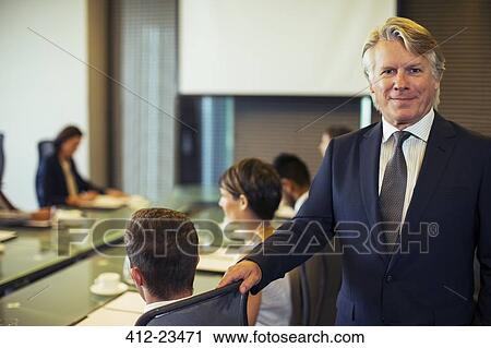 Stock Image - Portrait of businessman standing in conference room with colleagues in background. Fotosearch