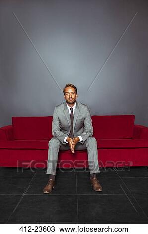 Stock Image - Portrait of businessman wearing grey suit sitting with hands clasped in dark room. Fotosearch