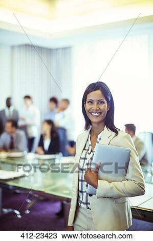 Stock Image - Portrait of businesswoman holding laptop in conference room. Fotosearch