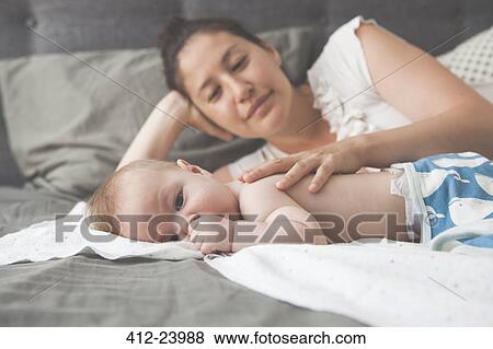 Stock Photo - Portrait of little baby sucking thumb with mother smiling in background. Fotosearch