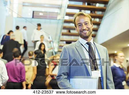 Portrait of smiling businessman holding file, standing in crowded lobby of conference center View Large Photo Image Stock Photo - Portrait of smiling businessman holding file, standing in crowded lobby of conference center. Fotosearch