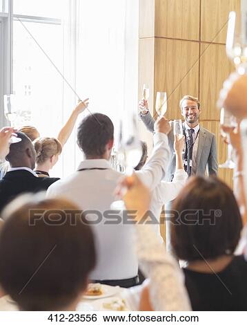 Stock Photograph - Portrait of smiling man standing before audience in conference room, raising toast with champagne flute. Fotosearch