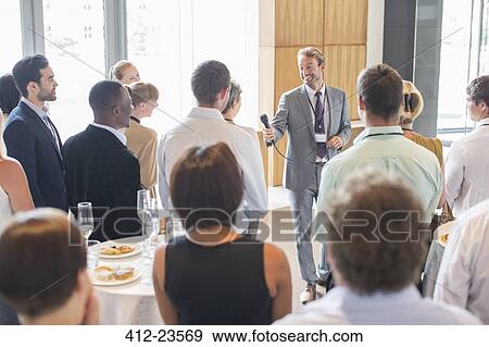 Stock Photo - Portrait of smiling man standing in conference room, offering microphone to person in audience. Fotosearch