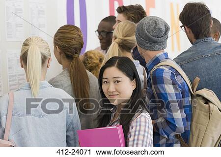 Stock Image - Portrait of smiling university student standing in corridor, people in background looking at exam results. Fotosearch