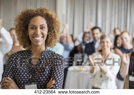 Portrait of smiling young woman in conference room with people applauding in background View Large Photo Image Picture - Portrait of smiling young woman in conference room with people applauding in background. Fotosearch