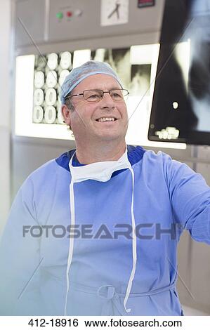 Stock Photograph - Smiling mature doctor wearing surgical clothing looking at x-ray in hospital. Fotosearch