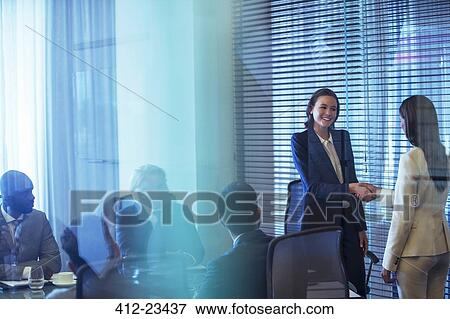 Stock Photo - Two businesswomen shaking hands in conference room, colleagues watching. Fotosearch