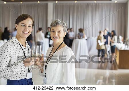 Stock Image - Two businesswomen standing with champagne flutes in hands, colleagues in background. Fotosearch