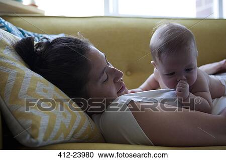 Stock Image - View of mother lying on sofa with little baby sucking thumb. Fotosearch