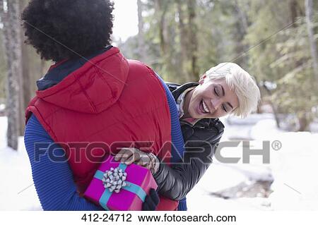 Stock Image - Woman peeking at Christmas gift behind man's back. Fotosearch