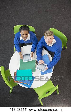 Stock Photo - Overhead view of teenage boy and girl sitting at round table with digital tablets, looking at camera. Fotosearch