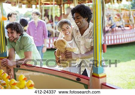 Stock Photo - Smiling couple holding teddy bear next to fishing game in amusement park. Fotosearch