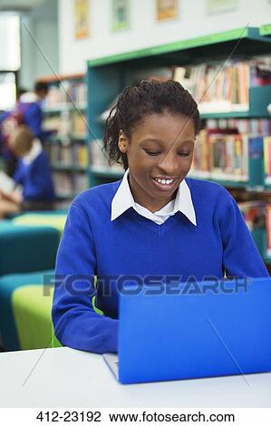 Stock Image - Smiling female student wearing blue school uniform using laptop in library. Fotosearch