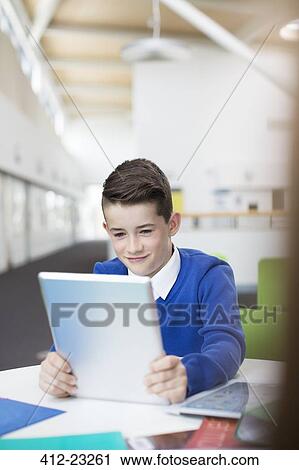 Stock Image - Smiling primary school boy wearing blue school uniform holding digital tablet at desk. Fotosearch