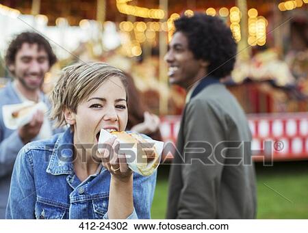 Stock Image - Three young people eating hot-dogs in amusement park. Fotosearch