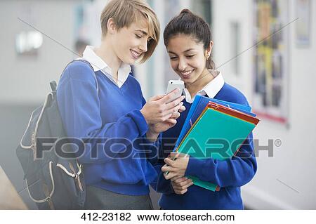 Stock Image - Two smiling female students wearing school uniforms looking at smartphone in school corridor. Fotosearch