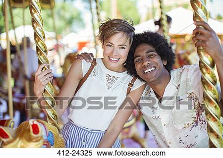 Stock Photography - Young multiracial couple smiling on carousel in amusement park. Fotosearch