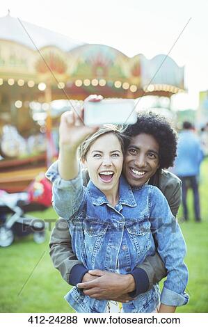 Stock Photo - Young multiracial couple taking selfie in amusement park. Fotosearch