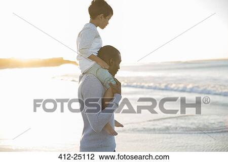Stock Image - Father carrying son on shoulders on beach. Fotosearch