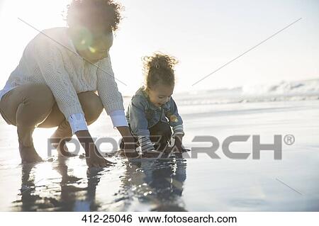 Stock Photograph - Mother and daughter crouching on beach and touching water. Fotosearch