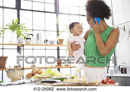 Mother holding her baby daughter while using mobile phone in kitchen View Large Photo Image Stock Image - Mother holding her baby daughter while using mobile phone in kitchen. Fotosearch