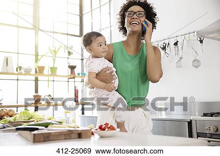 Stock Photo - Mother holding her baby daughter while using mobile phone in kitchen. Fotosearch