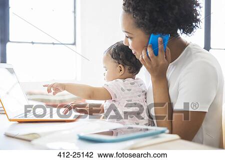 Stock Image - Mother working from home with daughter sitting on her lap. Fotosearch