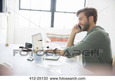 Stock Image - Portrait of young man sitting at desk with mobile phone and laptop. Fotosearch