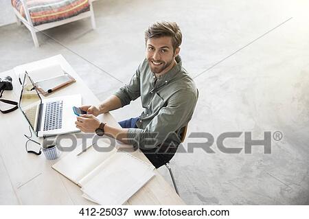 Stock Photo - Portrait of young man sitting at desk with mobile phone and laptop. Fotosearch