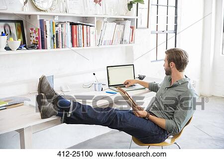 Stock Image - Portrait of young man sitting with legs on desk working with laptop. Fotosearch