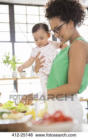 Stock Photograph - Woman with baby girl preparing meal in domestic kitchen. Fotosearch