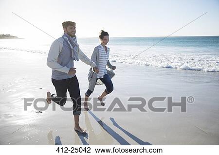 Stock Image - Young couple holding hands and running on beach. Fotosearch