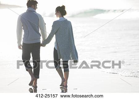 Stock Photo - Young couple holding hands and walking on beach. Fotosearch