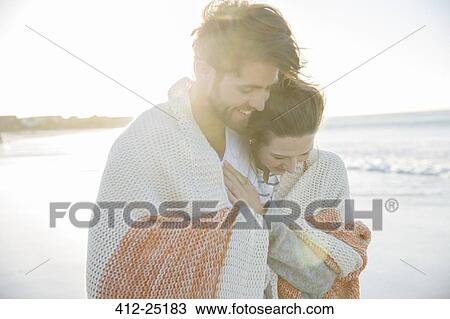 Stock Image - Young couple wrapped in blanket on beach. Fotosearch
