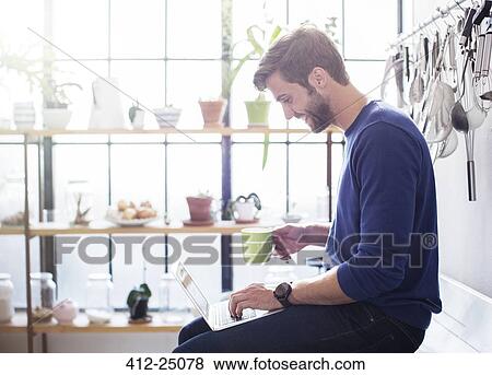 Stock Photo - Young man sitting on kitchen counter with mug and laptop. Fotosearch