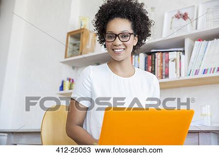 Stock Image - Young woman working with orange laptop in studio. Fotosearch