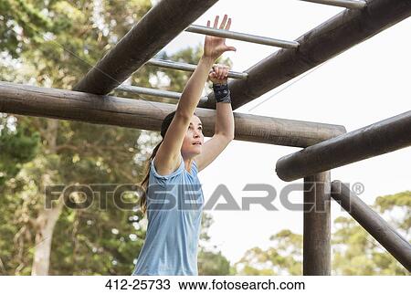 Stock Image - Determined woman crossing monkey bars on boot camp obstacle course. Fotosearch