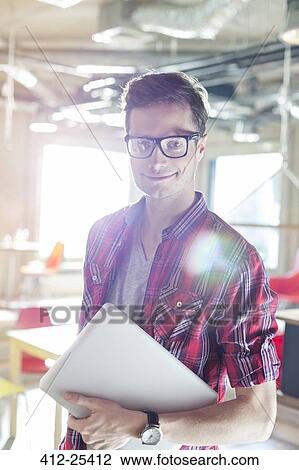 Stock Image - Portrait of casual businessman holding laptop in sunny office. Fotosearch