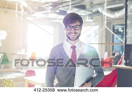 Portrait of confident businessman holding laptop in sunny office View Large Photo Image Stock Photo - Portrait of confident businessman holding laptop in sunny office. Fotosearch