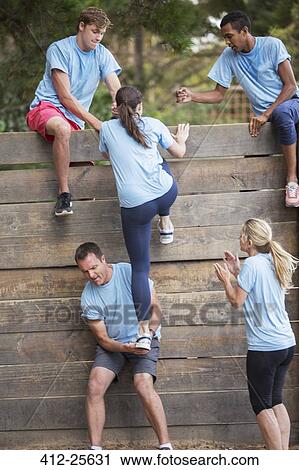 Stock Image - Teammates helping woman over wall on boot camp obstacle course. Fotosearch