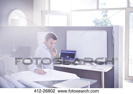 Stock Image - Architect reviewing blueprints in office. Fotosearch