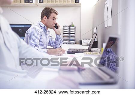 Picture - Architect talking on cell phone and working at laptop in office. Fotosearch