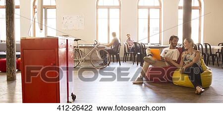 Stock Image - Casual business people working at laptop on bean bag chairs in open office. Fotosearch