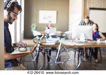 Casual businessman with headphones using laptop in office View Large Photo Image Stock Image - Casual businessman with headphones using laptop in office. Fotosearch